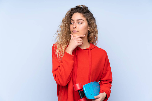 Young blonde woman with curly hair isolated on blue background with a skate and looking lateral - Fotó, kép