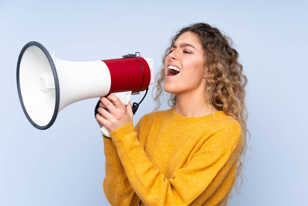 Young blonde woman with curly hair isolated on blue background shouting through a megaphone - Foto, imagen