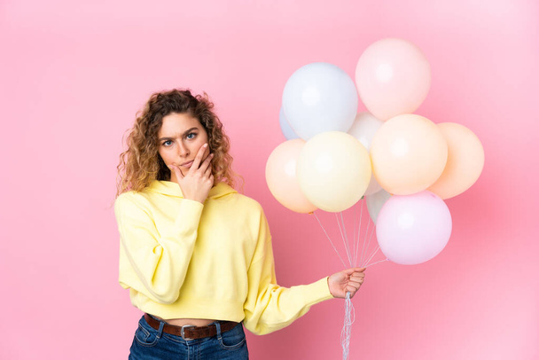 Jovem loira com cabelos cacheados pegando muitos balões isolados no fundo rosa pensando uma ideia - Foto, Imagem