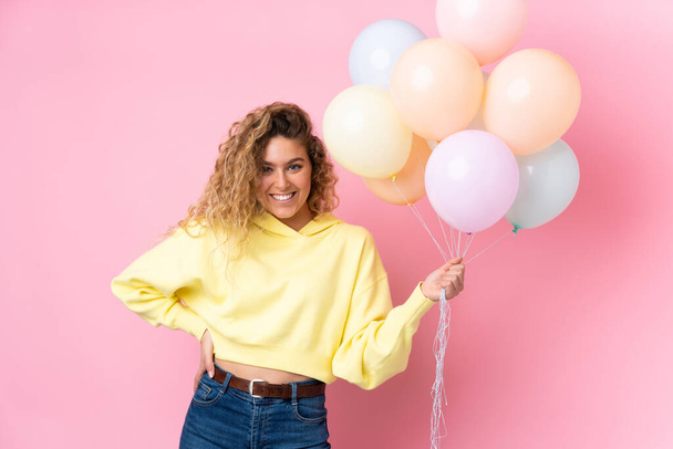 Young blonde woman with curly hair catching many balloons isolated on pink background - Foto, afbeelding