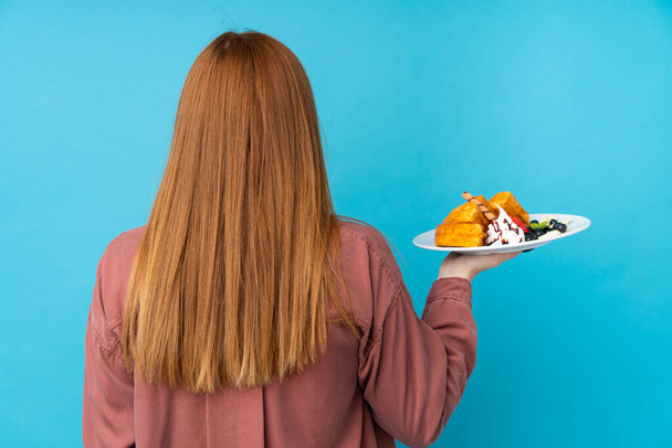 Genç kızıl saçlı kadın arka planda izole edilmiş arka planda waffle tutuyor. - Fotoğraf, Görsel