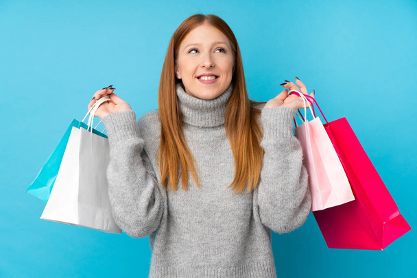 Young redhead woman over isolated blue background holding shopping bags and smiling - 写真・画像