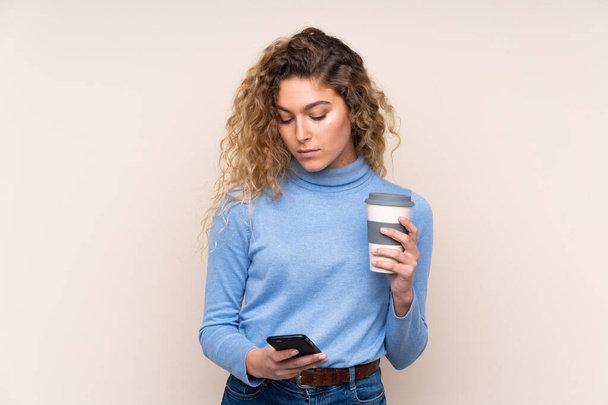 Молода блондинка з кучерявим волоссям у светрі з черепашкою ізольована на бежевому фоні, що тримає каву, щоб забрати і мобільний
 - Фото, зображення