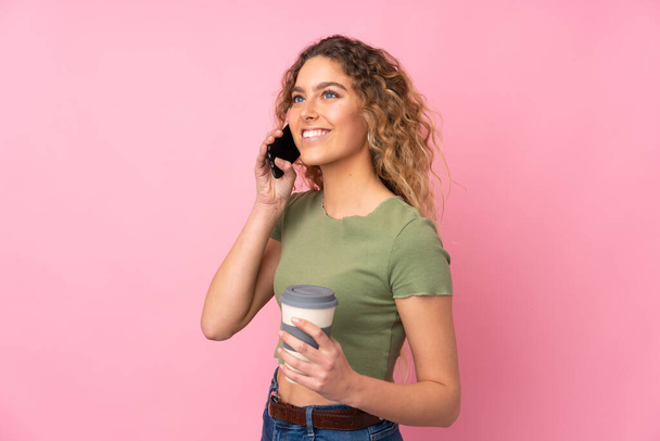 Junge blonde Frau mit lockigem Haar isoliert auf rosa Hintergrund hält Kaffee zum Mitnehmen und ein Handy - Foto, Bild