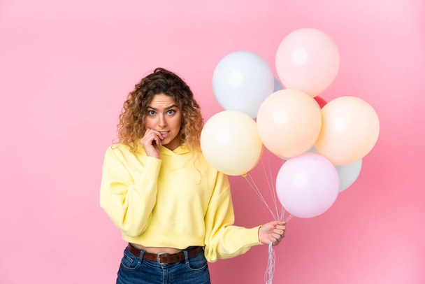 Young blonde woman with curly hair catching many balloons isolated on pink background nervous and scared - Foto, Imagem