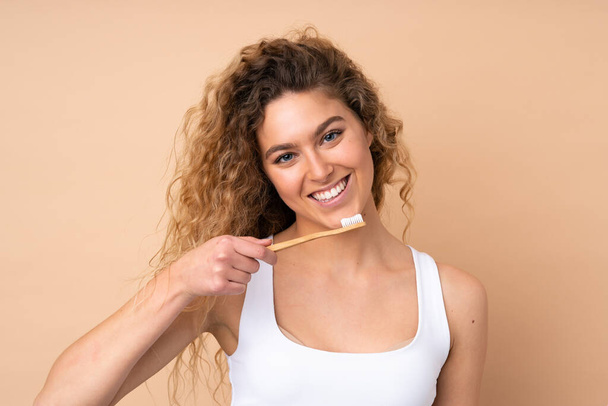 Νεαρή ξανθιά γυναίκα με σγουρά μαλλιά απομονωμένη σε μπεζ φόντο με οδοντόβουρτσα και χαρούμενη έκφραση - Φωτογραφία, εικόνα