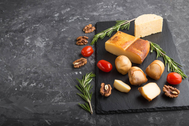 Smoked cheese and various types of cheese with rosemary and tomatoes on black slate board on a black concrete background. Side view, copy space, selective focus. - Photo, Image