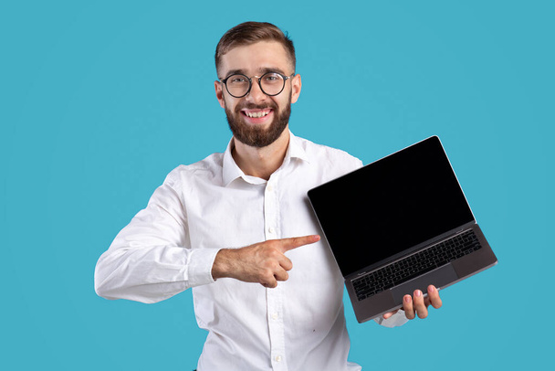 Happy millennial businessman pointing at laptop computer with mockup for website design on blue studio background - Photo, Image