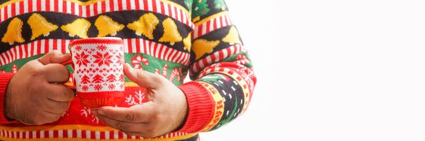 man drinking hot chocolate. Christmas tree pattern. Decorated drink mug. male wearing knitted sweater with Xmas ornament holding hot coffee in red cup. Gay celebrate winter holidays. no face - Foto, imagen