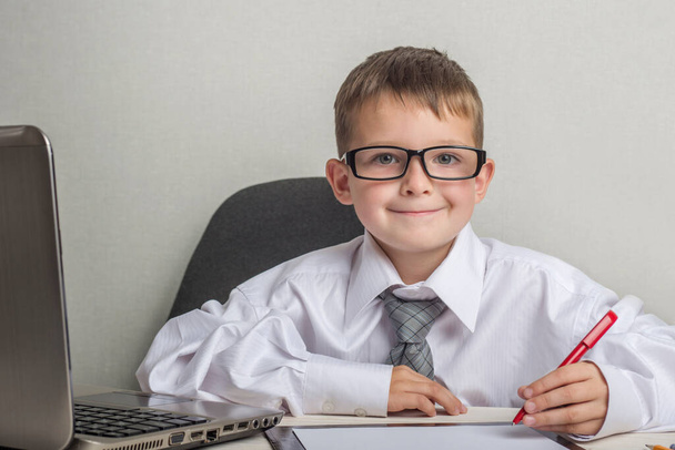 An adorable child is writing on a clipboard in suit and glasses. Little boss in the office. Children pretend to be adults. - 写真・画像