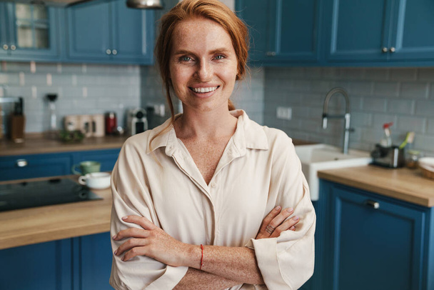 Image of happy redhead woman smiling and looking at camera while standing at home kitchen - Photo, image