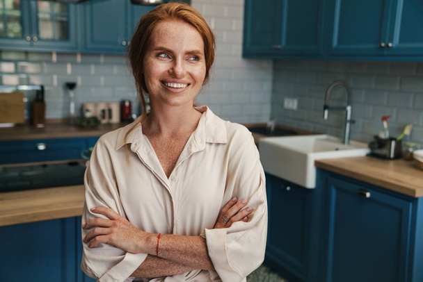 Image of happy redhead woman smiling and looking aside while standing at home kitchen - Foto, immagini