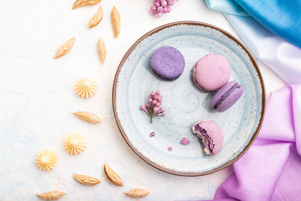 Purple macarons or macaroons cakes with cup of coffee on a white concrete background and magenta-blue textile. Top view, flat lay, close up. - Фото, изображение