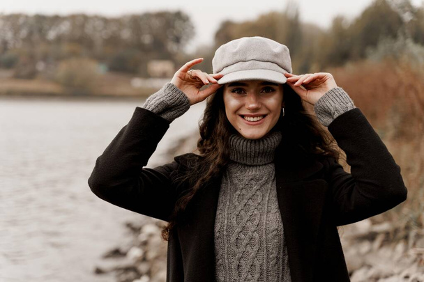 Красивая девушка в черном пальто и серой шерстяной кепке у озера. Путешествие по вашей стране - Фото, изображение