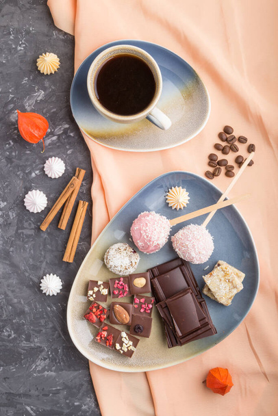 Ein Stück hausgemachte Schokolade mit Kokosbonbons und einer Tasse Kaffee auf schwarzem Betongrund und orangefarbenem Textil. Draufsicht, flach liegend, Nahaufnahme. - Foto, Bild