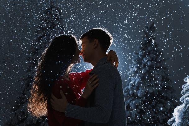 Vallende sneeuw en kussen met vallende sneeuw en donkerblauwe achtergrond. Gelukkig jong stel droeg witte truien. knuffels en kussen in de buurt van kerstbomen aan de vooravond van nieuwjaarsviering in de winterdag - Foto, afbeelding