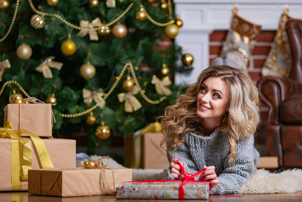 Nice blonde with christmas presents near new year tree. Luxury interior before holiday. - Фото, изображение