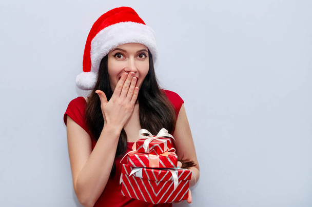 Κοντινό πλάνο Χριστουγεννιάτικη φωτογραφία του Εντυπωσιασμένο κορίτσι Screaming Wow OMG με κουτιά δώρων σε κόκκινο φόρεμα και Σάντα καπέλο - Φωτογραφία, εικόνα