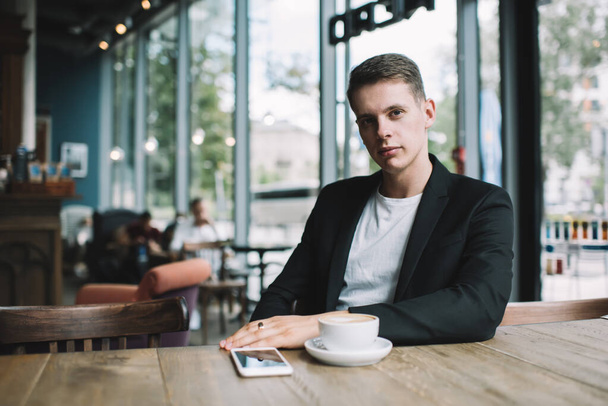 Young fair male in relaxed pose and looking at camera in cafe with full wall windows in daytime with coffee and smartphone on wooden table - 写真・画像