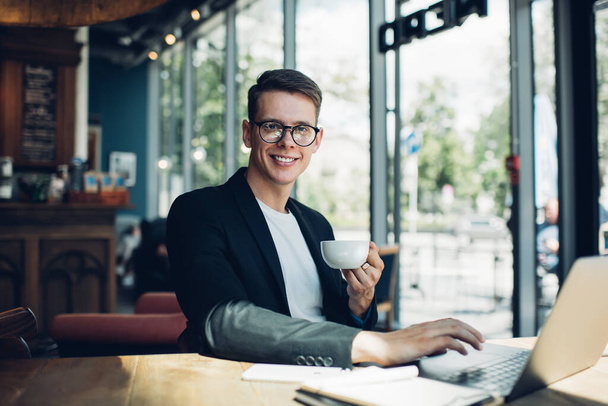 Positive young male in formal suit and eyeglasses sitting at table with cup of hot drink and laptop while working in cafe - Foto, Bild