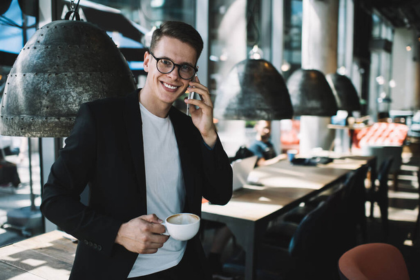 Young cheerful well dressed male in eyeglasses smiling and looking at camera while standing with cup of hot drink and speaking on mobile phone in cafe - Foto, imagen