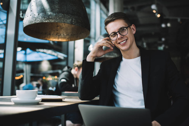 Smiling young man in formal suit and eyewear holding laptop and looking at camera while sitting at table in modern cafe - Foto, Bild