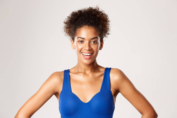 Крупним планом приваблива здорова жінка в синьому спортивному бюстгальтері, посміхаючись на камеру, стоїть над білим тлом
 - Фото, зображення