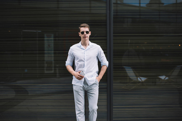 Trendy young male in casual outfit with sunglasses leaning on glass wall of modern building at daytime and holding smartphone - Фото, изображение