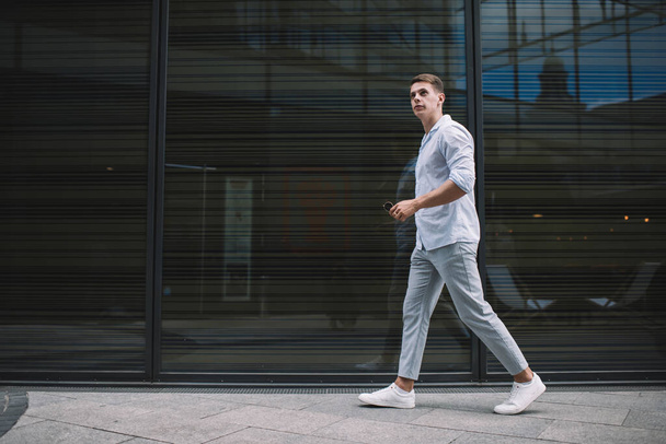 Young serious male in casual wear taking sunglasses while walking on sidewalk and passing by dark transparent wall in daylight and looking away - Photo, Image