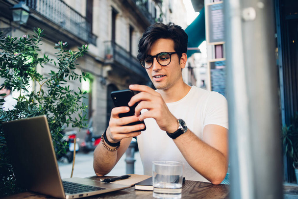 Positive stylish man in glasses and watch sitting at cafe on street of Barcelona with laptop and water on table while using smartphone - Fotó, kép