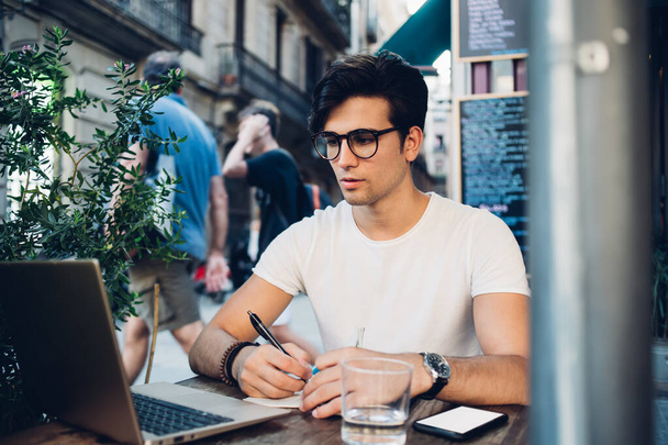 Serious male freelancer in eyeglasses and casual clothes sitting at table in street cafe and making notes in copybook while working on  laptop - Φωτογραφία, εικόνα