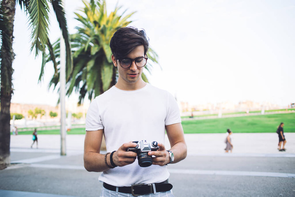 Positive caucasian hipster guy holding camera taking photos during journey exploring city destinations, handsome 20s pensive man tourist making pictures resting in town during summer vacations - Photo, Image