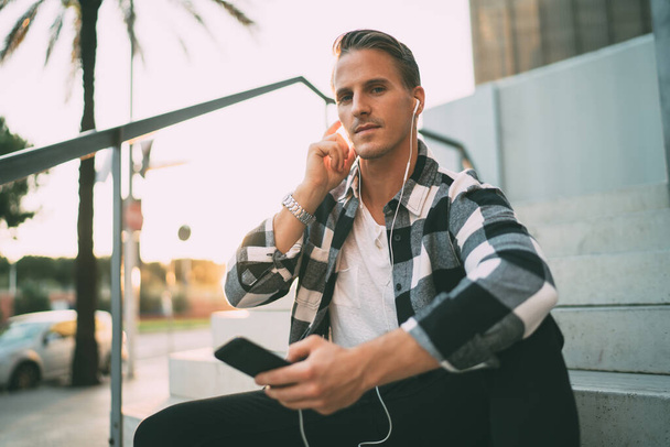 Portrait of handsome caucasian hipster guy in earphones listening music via playlist app on mobile phone sitting on street, pensive man looking at camera enjoying audiobook on smartphone in town - Photo, image