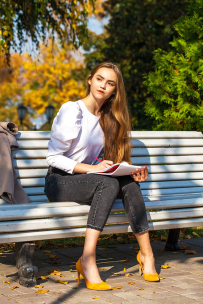 Young beautiful girl draws with a pencil in the sketchbook while sitting in the autumn park - Foto, Imagem
