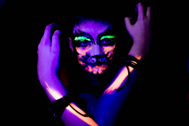 Le garçon a un visage peint d'un chat en colère dans des couleurs néon, rayonnant sous la lumière ultraviolette. - Photo, image
