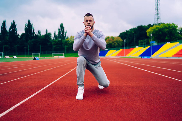 Young concentrated bearded male in sportswear and sneakers stretching legs and warming up while preparing for training on empty running track - Foto, Imagem