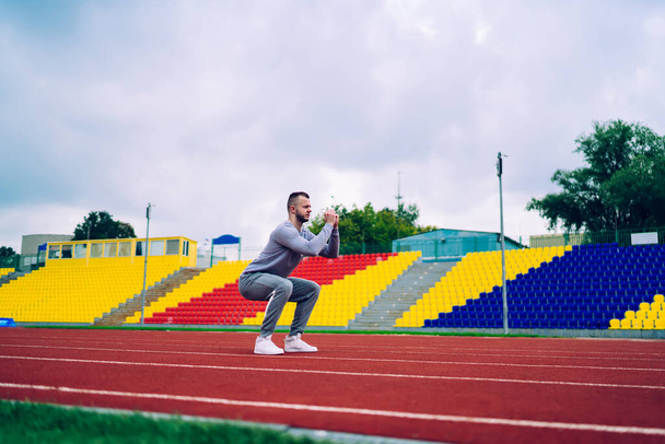 Full body of focused young bearded guy in active wear doing squats on stadium track with empty tribune during intensive workout on cloudy day - Фото, зображення