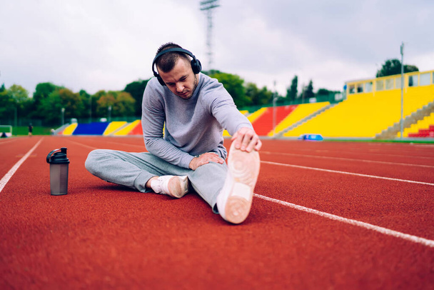 Serious young athletic male in sportswear listening to music in headphones and drinking water while resting and stretching legs after workout on stadium - Foto, Bild