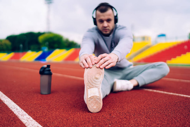Selective focus on high quality sneakers sole for running and training,blurred young strong caucasian man in headphones stretching lower body muscles and listening music on stadium on workout - Foto, imagen