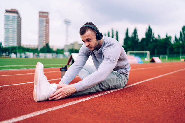 Serious male athlete in sportswear and sneakers sitting on running track and stretching legs before training while listening to music - Foto, Imagem
