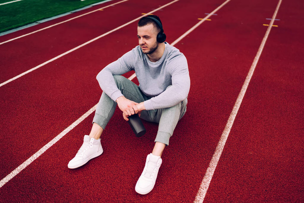From above of full length sportsman in activewear and sneakers sitting on track and listening to music in headphones while resting after training - Foto, imagen
