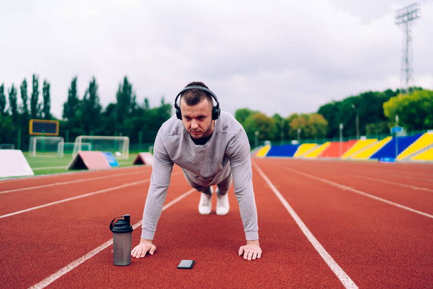 Strong determined caucasian male keeping healthy physical strength on training listening music online via headphones,20s serious sportsman in earphones standing in plank having workout on stadium - Foto, Imagem