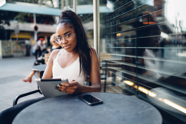 Beautiful dark skinned young hipster girl in trendy eyewear looking at camera holding tablet for networking on cafe terrace, portrait of smiling african american woman typing message on touchpad - Fotó, kép