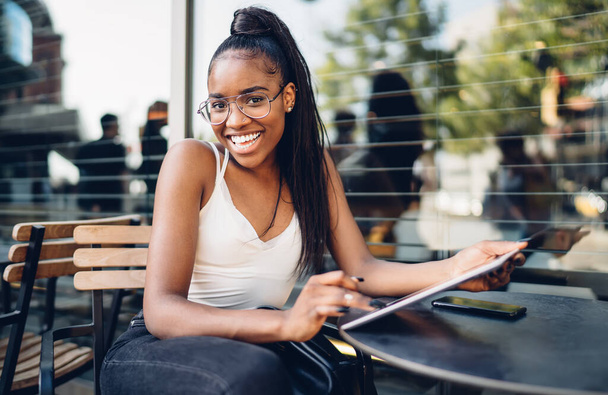 Portrait of cheerful dark skinned woman blogger satisfied with 4G internet connection and sharing in social networks, smiling african american female millennial looking at camera making online booking - Foto, afbeelding