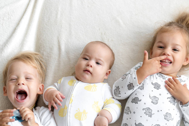 Трое маленьких улыбающихся детей играют вместе на кровати. Брат и сестра показывают новорожденному игрушку. Дети знакомятся с новорожденными братьями и сестрами. Тоддлеры смеются и сближаются. Смешные дети с небольшой разницей в возрасте. - Фото, изображение