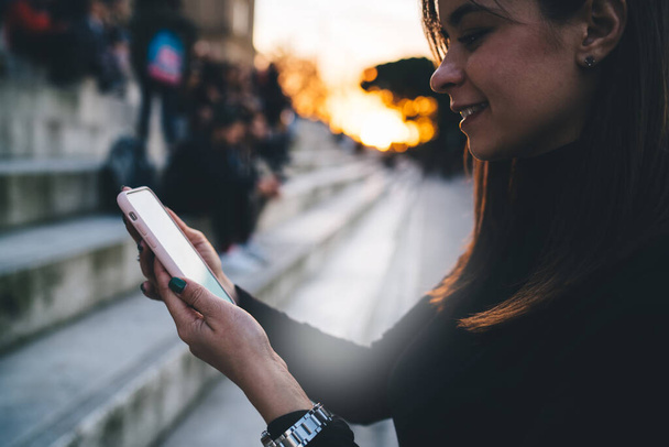 Side view of smiling young woman in casual clothes and wristwatch reading messages on smartphone while standing on street near steps - Photo, image