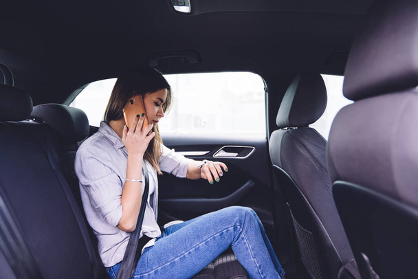 Side view of worried young female in hurry talking on smartphone and checking time on wristwatch while sitting in passenger seat while stuck in traffic - Foto, imagen