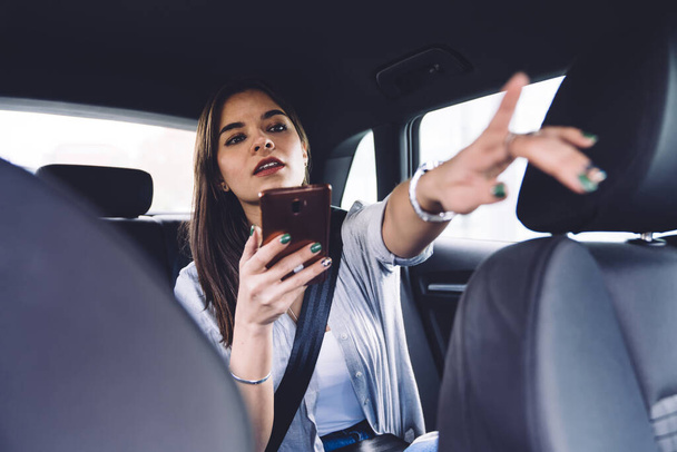 Young female passenger in casual clothing with smartphone sitting in comfortable backseat of cab giving direction to driver and pointing with finger - Photo, image