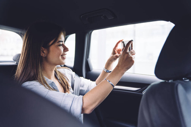 Side view of positive young brunette in casual wear smiling while riding on backseat of car and taking photo on mobile phone - Foto, Bild
