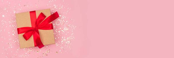 Slavnostní prapor - dárková krabička s červenou mašlí na slavnostním růžovém pozadí se zlatými jiskřičkami kolem a kopírovacím prostorem. Plochý styl. Vánoce, Nový rok, Valentýn a narozeninová oslava koncepce - Fotografie, Obrázek
